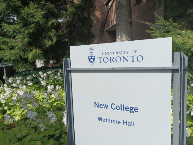 Außenansicht des neuen Colleges der Universität Toronto an der Wetmore Hall, mit einem Schild vor dem Gebäude, umgeben von grünem Gras, bunten Blumen, hohen Bäumen und einem hellblauen Himmel.