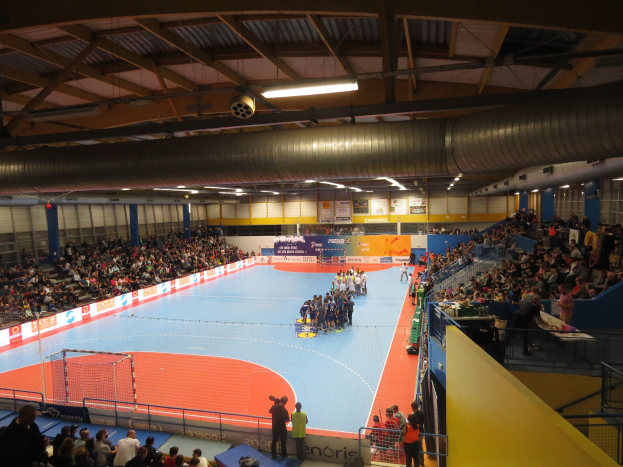 Großer Hallenstadium mit Zuschauern bei einem Handballspiel, mit Sitzplätzen, Stehplätzen, Netz auf der linken Seite, Hintergrundtafeln mit Text und Deckenbeleuchtung und Rohren.