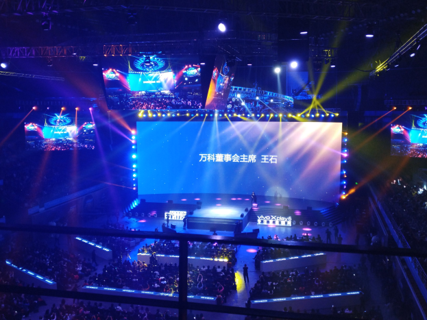 Eine große Menschenmenge sitzt vor einer hell erleuchteten Bühne mit Metallabsperrungen und wartet auf ein Esport-Event mit einem großen Bildschirm im Hintergrund.