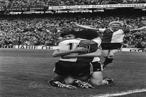 Drei Fussballspieler umarmen sich auf dem Boden nach einem Tor, mit jubelnder Menge und schwenkenden Fahnen im Hintergrund, in einem schwarz-weiss gestalteten Stadion.