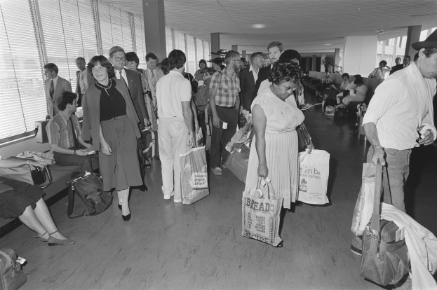 Ein Schwarz-Weiß-Bild von Reisenden in einem Flughafen, einige sitzen auf Bänken und andere stehen, mit einem Mann und einer Frau, die Taschen halten, in der Mitte und Jalousien auf der linken Seite.