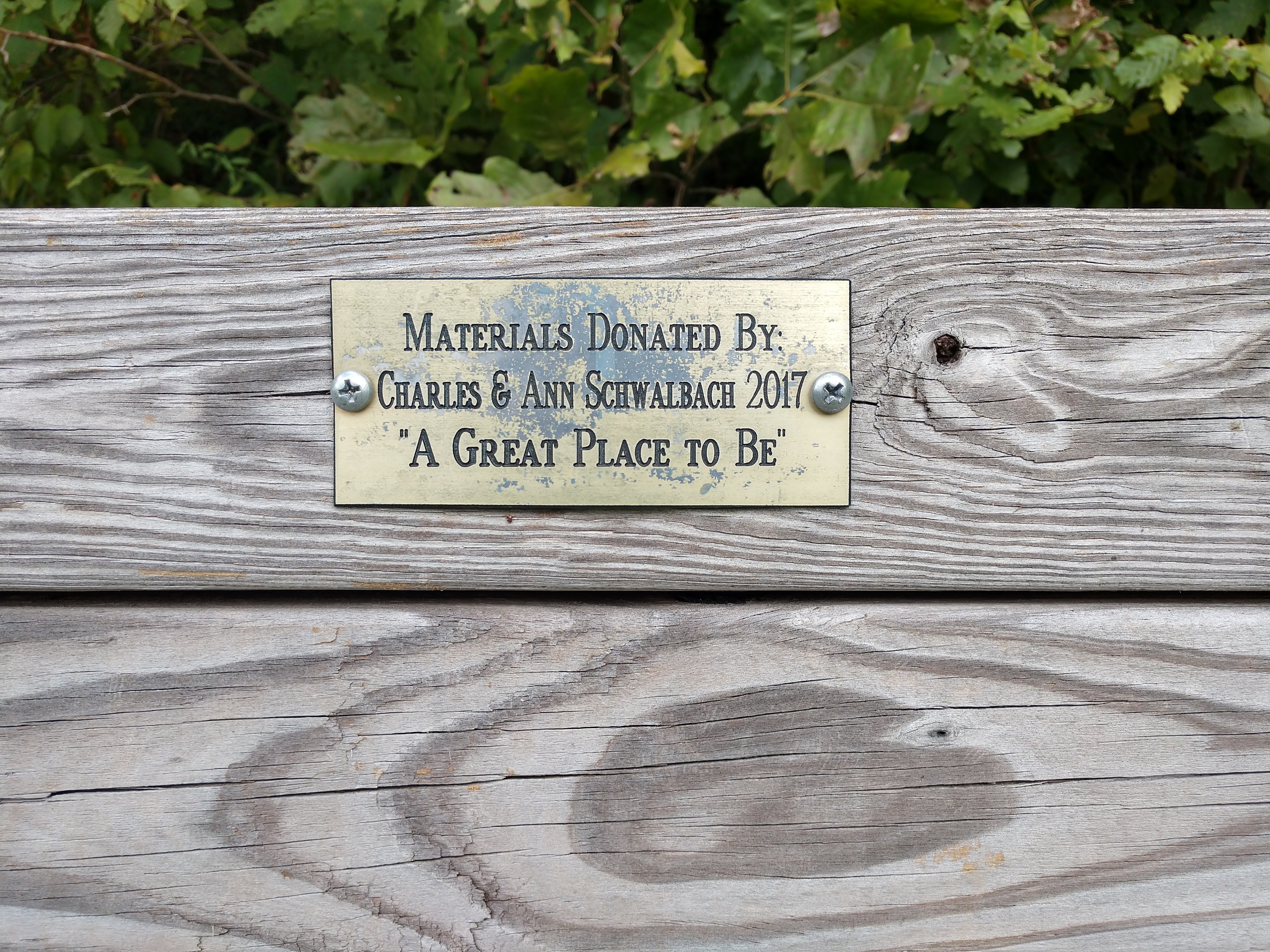 Holzbank mit einer Plakette mit der Aufschrift "Materialien gespendet von Charles & Ann Schwabach 2017: Ein toller Ort zu sein" in einem Garten mit üppiger grüner Vegetation.