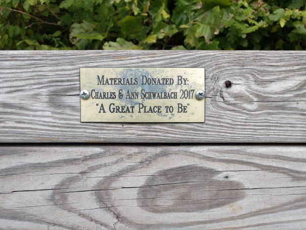 Holzbank mit einer Plakette mit der Aufschrift "Materialien gespendet von Charles & Ann Schwabach 2017: Ein toller Ort zu sein" in einem Garten mit üppiger grüner Vegetation.