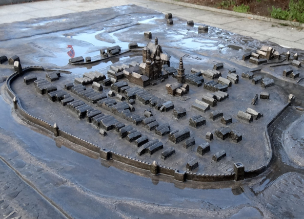 Eine Miniaturstadtmodell in einer Pfütze umgeben von Gebäuden, Bäumen, Pflanzen und einer Hintergrundwand.