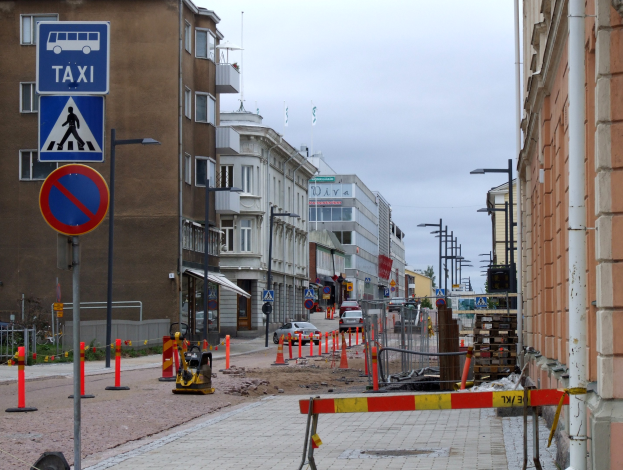 Stadtstraßenszene mit Gebäuden, Straßeninfrastruktur, Fahrzeugen, Bäumen und einer Baustelle mit Verkehrsschildern.