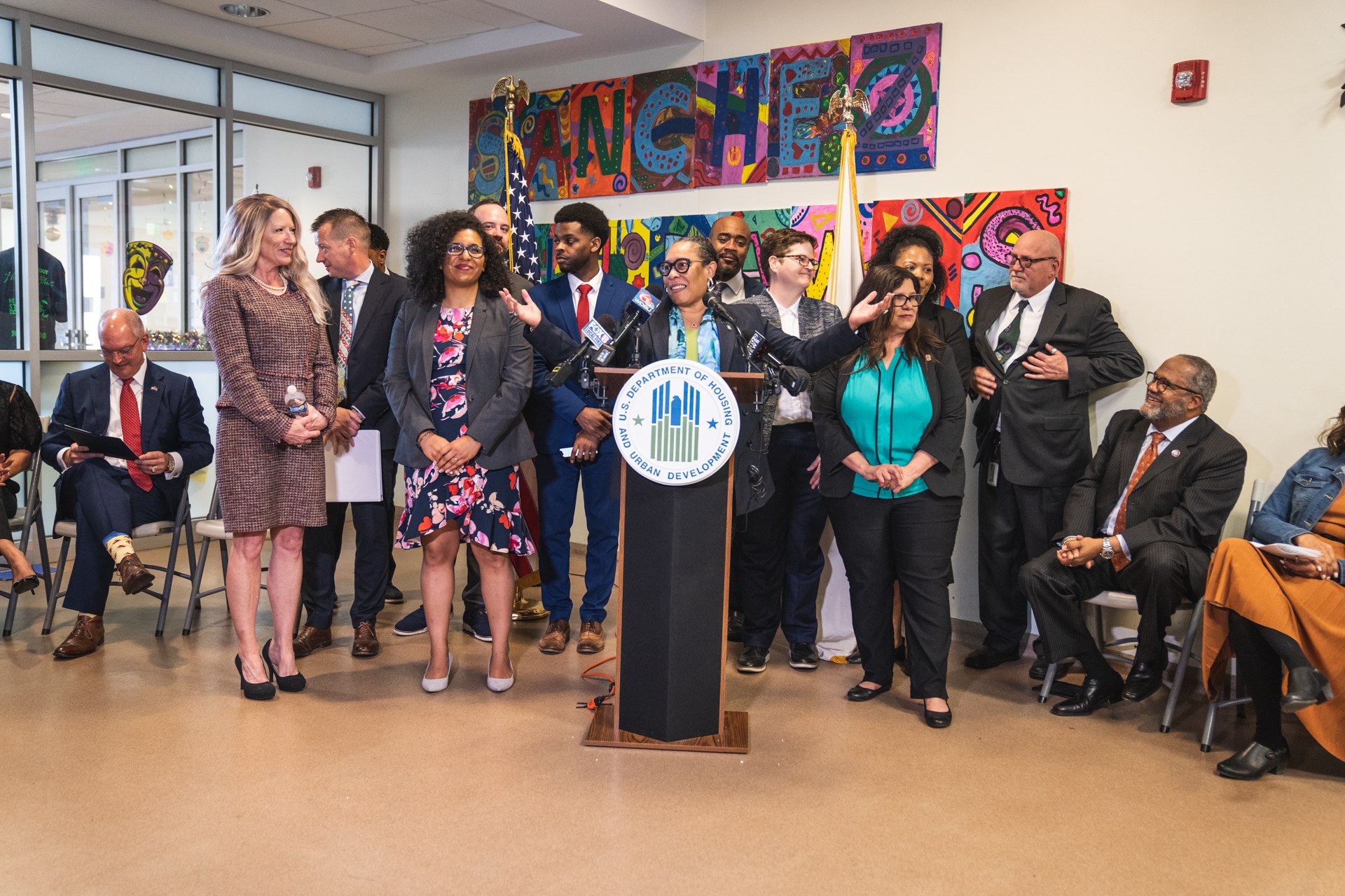 Gruppe von Menschen vor einem Rednerpult mit Mikrofon und einer Tafel mit der Aufschrift "Sancho City Councilman Joins City Council", mit sitzenden und stehenden Zuschauern, Fahnen, Feuerlöscher und Deckenleuchten im Hintergrund.