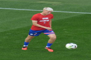 Ein junger Mann in einem roten Trikot und blauen Shorts, der Fußball auf einem grünen Feld spielt, trägt Fußballschuhe und -bekleidung während eines Spiels.
