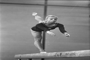 Eine Frau in einem Einteiler führt Gymnastik auf einem Balancebalken aus, ihre Arme sind ausgestreckt und ihre Beine bilden einen anmutigen Bogen, mit einem konzentrierten Gesichtsausdruck.