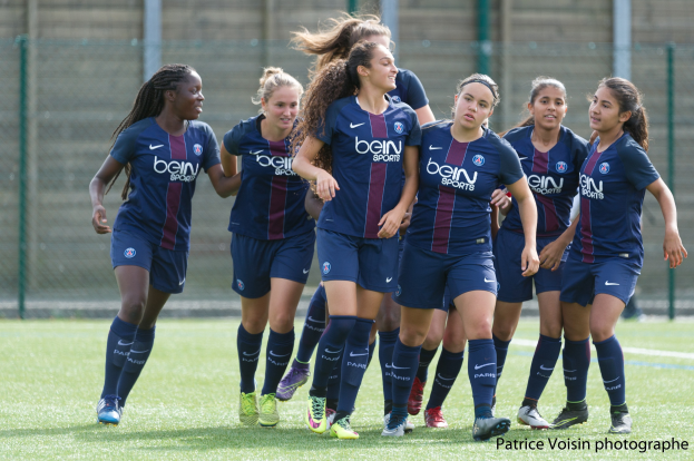 Eine Gruppe junger Frauen, die auf einem Rasenfeld Fußball spielen, mit Maschendrahtzaun und einer Wand im Hintergrund und dem Text "Paris Saint-Germain Frauenfußball" in der rechten unteren Ecke.