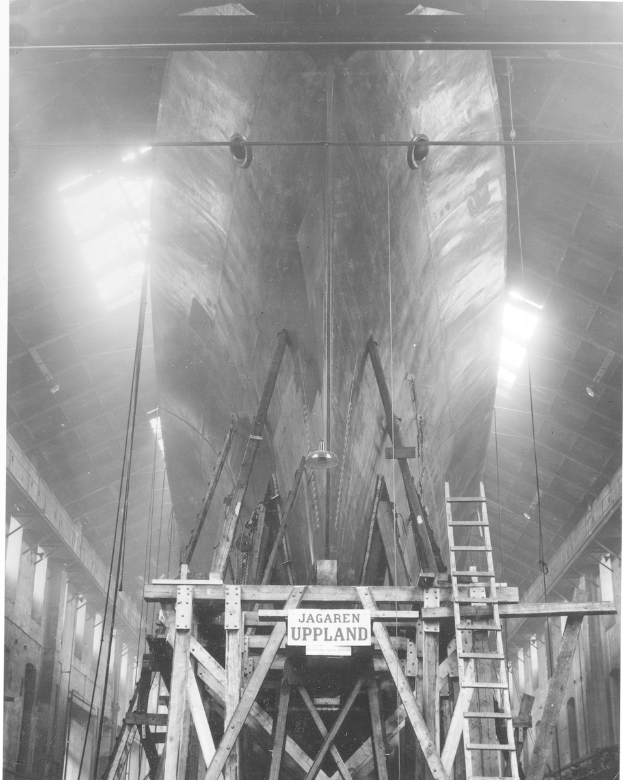Ein großes Boot im Bau in einer Werft, mit einer Leiter, Holzpfählen, einer Texttafel, Metallrahmen, Deckenlampen und Fenstern.