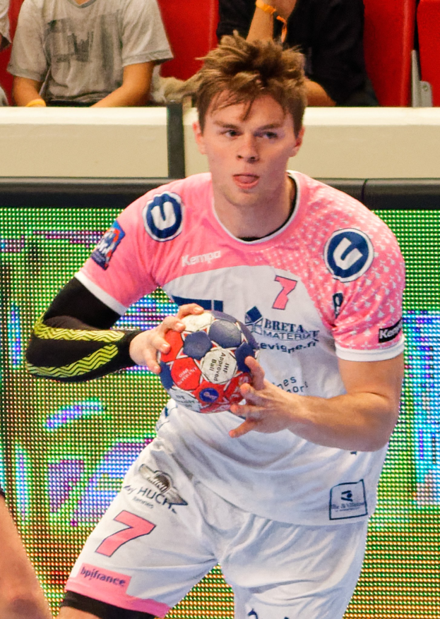 Ein Mann in einem pinken Hemd mit einem Fußball, daneben eine andere Person mit der Hand und im Hintergrund sitzende Menschen.
