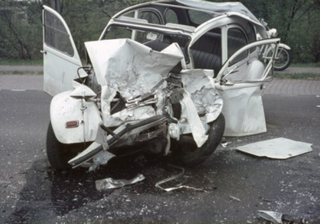 Ein schwer beschädigtwagen mit zertrümmertem Frontteil steht am Straßenrand, mit Bäumen und einem Gebäude im Hintergrund und ein weiteres Fahrzeug in der Ferne sichtbar.
