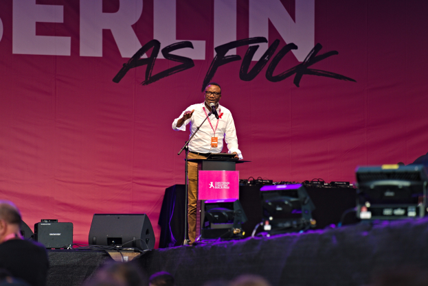 Ein Mann in einem weißen Hemd und braunen Hosen steht auf einer Bühne und hält ein Mikrofon in der Hand, während er eine Menschenmenge anspricht. Im Hintergrund ist ein pinkfarbenes Banner mit der Aufschrift "Berlin as Fuck" zu sehen.