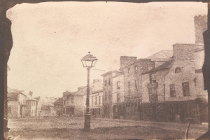 Altes Schwarz-Weiß-Foto einer Straßenlaterne, die in der Mitte einer Stadtstraße steht und von Gebäuden flankiert wird.