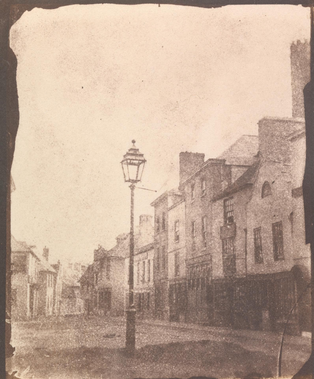 Altes Schwarz-Weiß-Foto einer Straßenlaterne, die in der Mitte einer Stadtstraße steht und von Gebäuden flankiert wird.