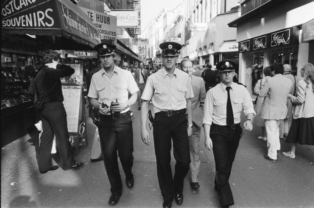 Drei Polizisten in Uniform gehen eine belebte Stadtstraße entlang, die von hohen Gebäuden, Geschäften und Passanten gesäumt ist.