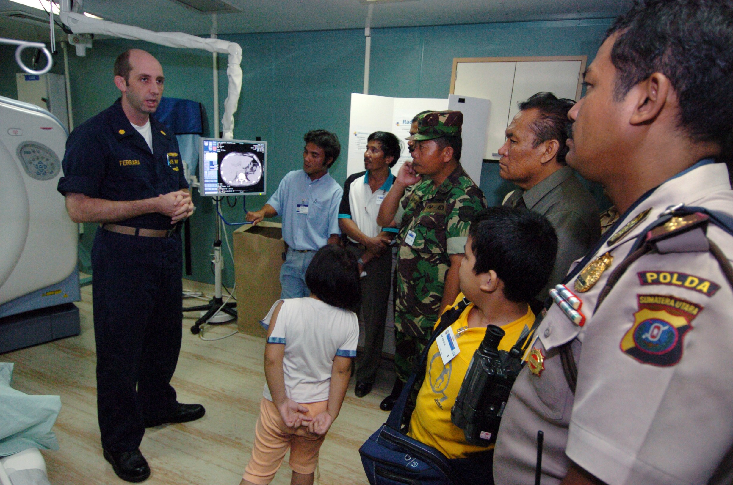 Eine Gruppe von Menschen in einem Krankenhauszimmer mit einem Mann, der ein Mikrofon hält, einem drapierten Bett auf der linken Seite, medizinischer Ausrüstung, einem Röntgengerät, einer Texttafel, einer Tür und Deckenleuchten.