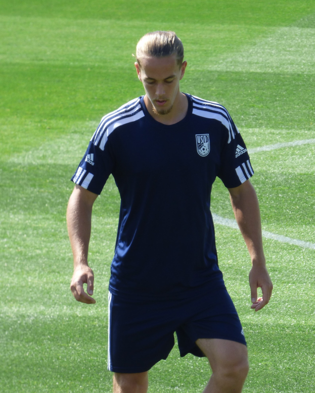 Ein professioneller Fußballspieler in einem blauen T-Shirt und Shorts geht selbstbewusst über ein grünes Feld.