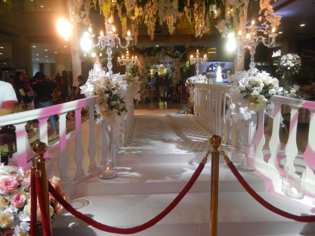 Eine Hochzeitszeremonie in einem Einkaufszentrum mit weißem Teppich, dekorativen Geländern, Blumenarrangements, Kronleuchtern und Deckenleuchten, bei der eine Menge Menschen mit Taschen anwesend ist.