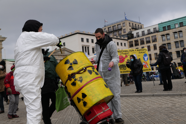 Eine Gruppe von Menschen in weißen Anzügen und Masken steht um einen gelben Eimer mit einem Wagen im Vordergrund, mit Schildern in der Hand, vor Gebäuden mit Fenstern und Fahnen an Stangen unter einem bewölkten Himmel.