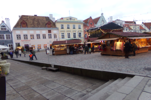Ein lebendiger Weihnachtsmarkt in Tallinn, Estland, mit Menschen um geschmückte Stände, festlicher Beleuchtung und Gebäuden mit Fenstern im Hintergrund, unter einem bewölkten Himmel, mit Treppen, die zum Markt führen, und Töpfen mit Pflanzen, die herumstehen.
