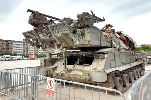 Ein gepanzerter Transporter steht vor einem Metallzaun mit einem daran befestigten Schild, mit Fahrzeugen, Bäumen, Gebäuden und einem klaren blauen Himmel im Hintergrund.