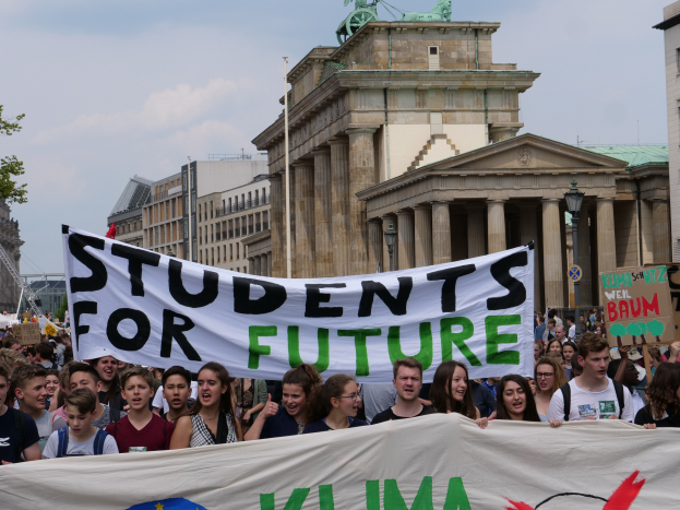 Eine Gruppe von Schülern marschiert in Berlin, hält eine bunt bemalte 'Students for Future'-Schriftzug-Tafel vor einer Kulisse aus Gebäuden, Bäumen und Himmel.