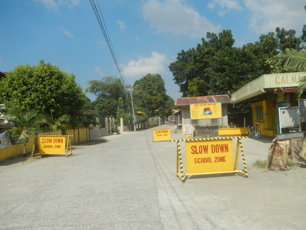 Gelbes "Langsam durch Schulzone"-Schild auf der linken Seite einer Straße mit parkenden Fahrzeugen auf der rechten Seite, Bäumen, Gebäuden, Strommasten und Drähten im Hintergrund.