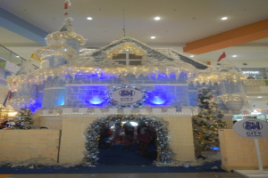 Ein festlich geschmücktes Weihnachtsdorf mit Burgenähnlicher Struktur, umgeben von Menschen und einem Textschild auf der rechten Seite, unter einer mit Lichtern geschmückten Decke in einem Einkaufszentrum.