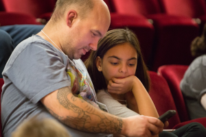 Zwei Menschen in einem Kino, ein Mann hält ein Handy, während ein junges Mädchen darauf schaut, mit anderen Sitzenden in der Nähe.
