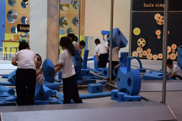 Kinder spielen in einer Turnhalle, einige halten Gegenstände, eine Wandtafel mit Text im Hintergrund und Stangen auf der rechten Seite.