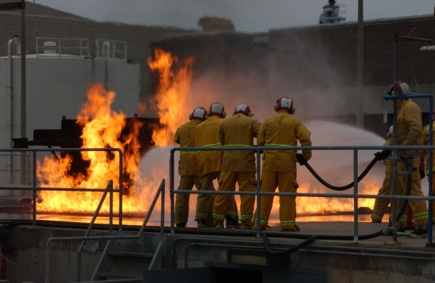 Firefighters in helmets stand on a building roof holding pipes, with railings and steps below and a building and sky in the background.