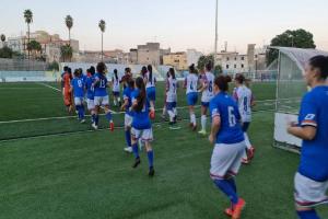 Eine Gruppe von Frauen, die auf einem Feld Fußball spielen, umgeben von Gebäuden, Bäumen und Pfählen unter einem klaren blauen Himmel, mit einem Torpfosten und Netz auf der rechten Seite.