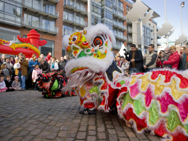 Ein lebendiges chinesisches Neujahrsfest in Amsterdam mit einer Drachenvorführung vor einer Menschenmenge, einige halten Kameras, vor einem Hintergrund aus Gebäuden, Laternenmästen und einem klaren blauen Himmel.