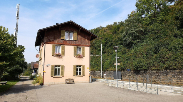 Ferienhaus zum Verkauf in Baden-Württemberg, Deutschland, mit einem Gebäude mit zahlreichen Fenstern, einer vorderen Mauer, umliegenden Straßen-Elementen, Grünflächen und einer bewölkten Himmel.