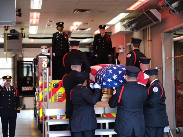 Eine Gruppe uniformierter Männer trägt einen Sarg mit einer amerikanischen Flagge, umgeben von einigen Männern mit Mützen und einem Feuerwehrauto sowie Deckenlichtern im Hintergrund.