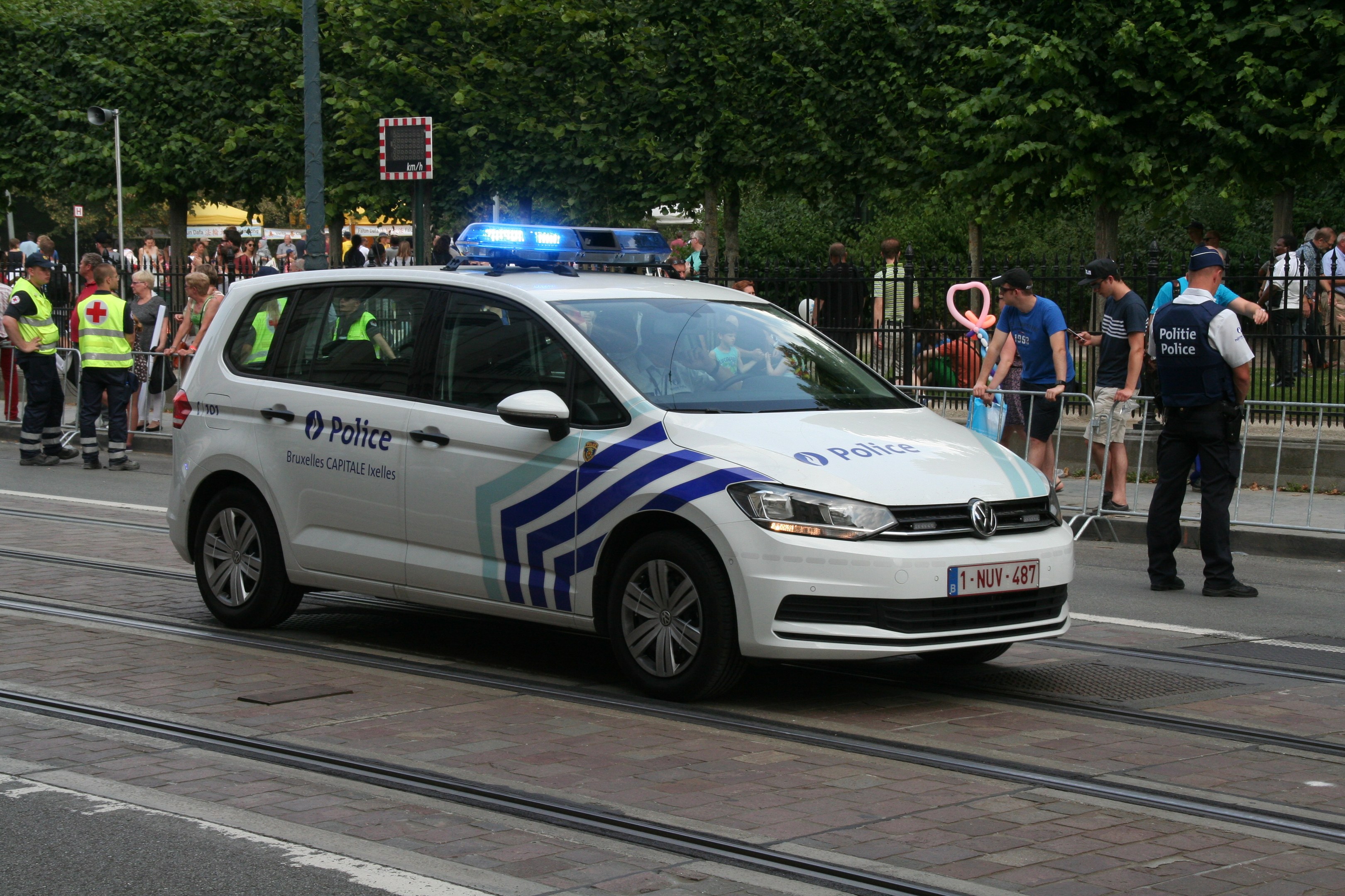 Polizeiauto fährt auf einer Straße neben einer Menschenmenge, einige tragen Mützen, mit einem Geländer, Bäumen und Laternen im Hintergrund.