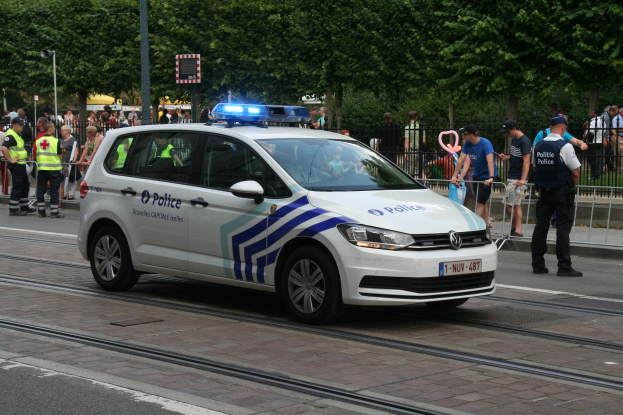 Polizeiauto fährt auf einer Straße neben einer Menschenmenge, einige tragen Mützen, mit einem Geländer, Bäumen und Laternen im Hintergrund.