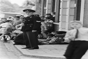 Ein Polizist steht vor einer Gruppe von Kindern, mit einem Gebäude auf der rechten Seite und Bäumen und Himmel im Hintergrund, dargestellt in Schwarz-Weiß.