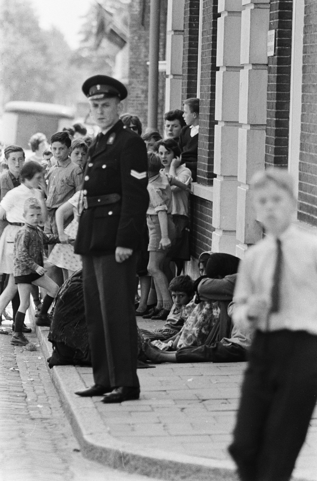 Ein Polizist steht vor einer Gruppe von Kindern, mit einem Gebäude auf der rechten Seite und Bäumen und Himmel im Hintergrund, dargestellt in Schwarz-Weiß.