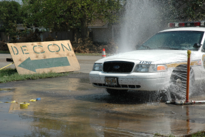 Ein Polizeiauto fährt durch eine Pfütze, mit einem Schild, einer Verkehrskegel und anderen Gegenständen am Boden, mit Bäumen, Gebäuden und einem klaren blauen Himmel im Hintergrund.