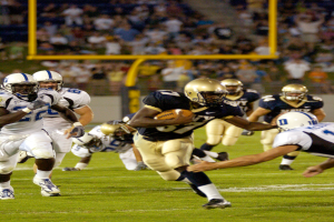 Ein Fußballspieler in Sportuniform und Helm rennt mit dem Ball, während er von einer Gruppe ähnlich gekleideter Spieler verfolgt wird, mit einem unscharfen Hintergrund und Text am unteren Bildrand.