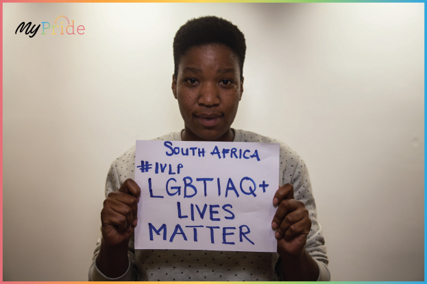 Eine Frau in einem weißen Hemd steht entschlossen vor einer Wand und hält ein Schild mit der Aufschrift "LGBTQ+ Lives Matter" in fetter schwarzer Schrift.