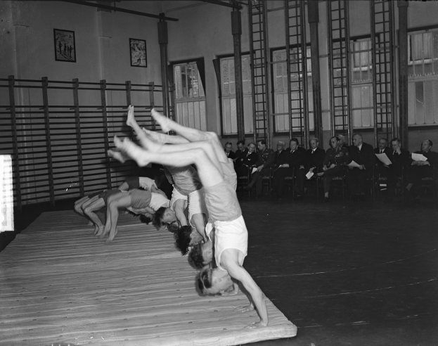 Eine Gruppe von Menschen führt Akrobatik auf einer Bühne vor einem sitzenden Publikum auf, mit Fotorahmen, Fenstern und Leitern im Hintergrund, dargestellt in Schwarz-Weiß.