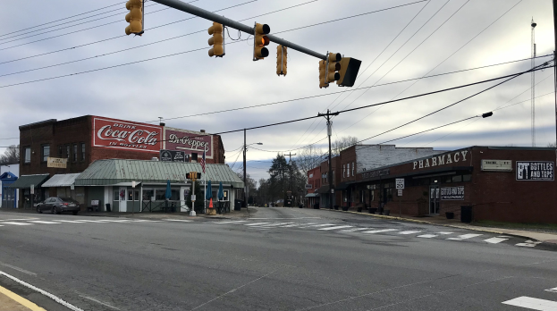 Stadtstraße mit Ampel, Fahrzeugen, Gebäuden mit Namensschildern, Strommasten mit Kabeln, Bäumen und einem bewölkten Himmel im Hintergrund.