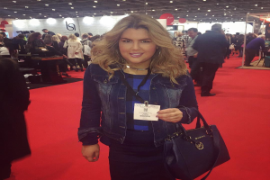 Eine Frau steht auf einem roten Teppich, lächelt und hält eine Handtasche in der Hand, umgeben von Menschen, Lichtern und Schildern auf einer Modenschau.