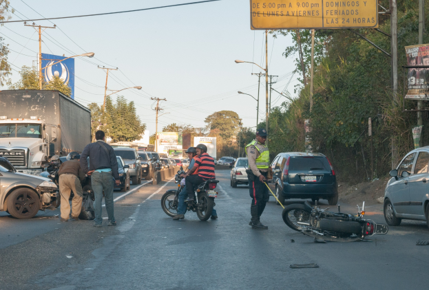 Gruppe von Menschen um ein verunglücktes Motorrad auf der Straße mit mehreren Fahrzeugen, Bäumen, Strommasten und Lichtern im Hintergrund.