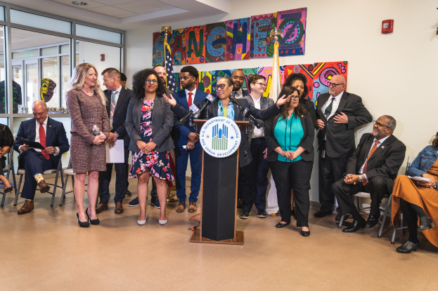 Eine Gruppe von Menschen, die vor einem Podium mit Mikrofon und einer Tafel mit der Aufschrift "Sancho-Stadtrat tritt dem Stadtrat bei" steht, mit Flaggen, einer Tafel, einem Feuerlöscher und Deckenleuchten an der Rückwand.