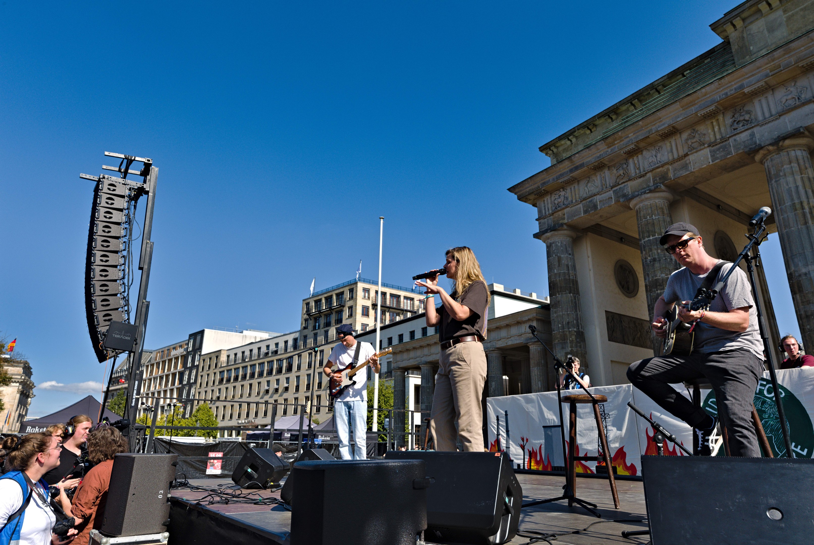 Gruppe von Menschen, die auf einer Bühne mit dem Brandenburger Tor im Hintergrund Musik machen, begleitet von Lautsprechern und Equipment unter einem klaren Himmel.