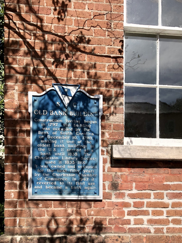 Altes Bankgebäude mit blauer Tafel an der Seite, einem Fenster rechts und einem Baum links; die Tafel enthält Text.